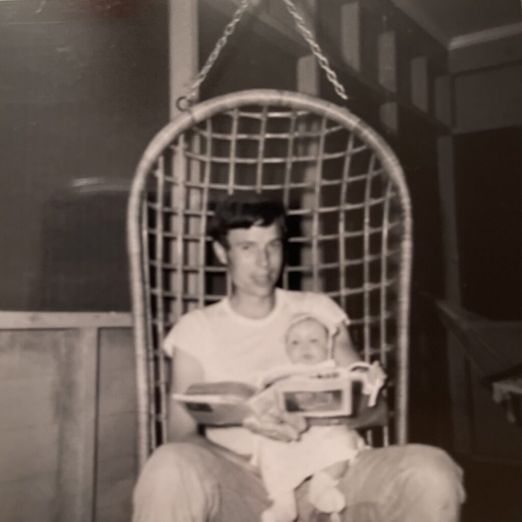 Dad sitting in a chair with me as a baby with a book reading.